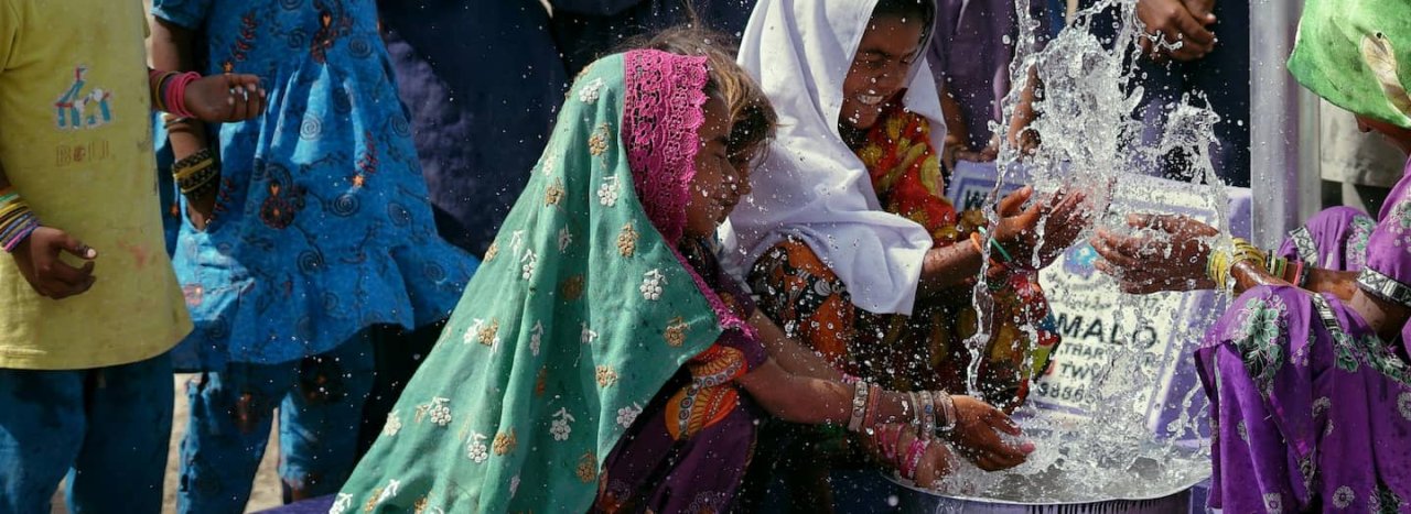 A fountain releasing water with a few ladies gathered round it, while children look on, representing courses in Transformational Development at All Nations college.